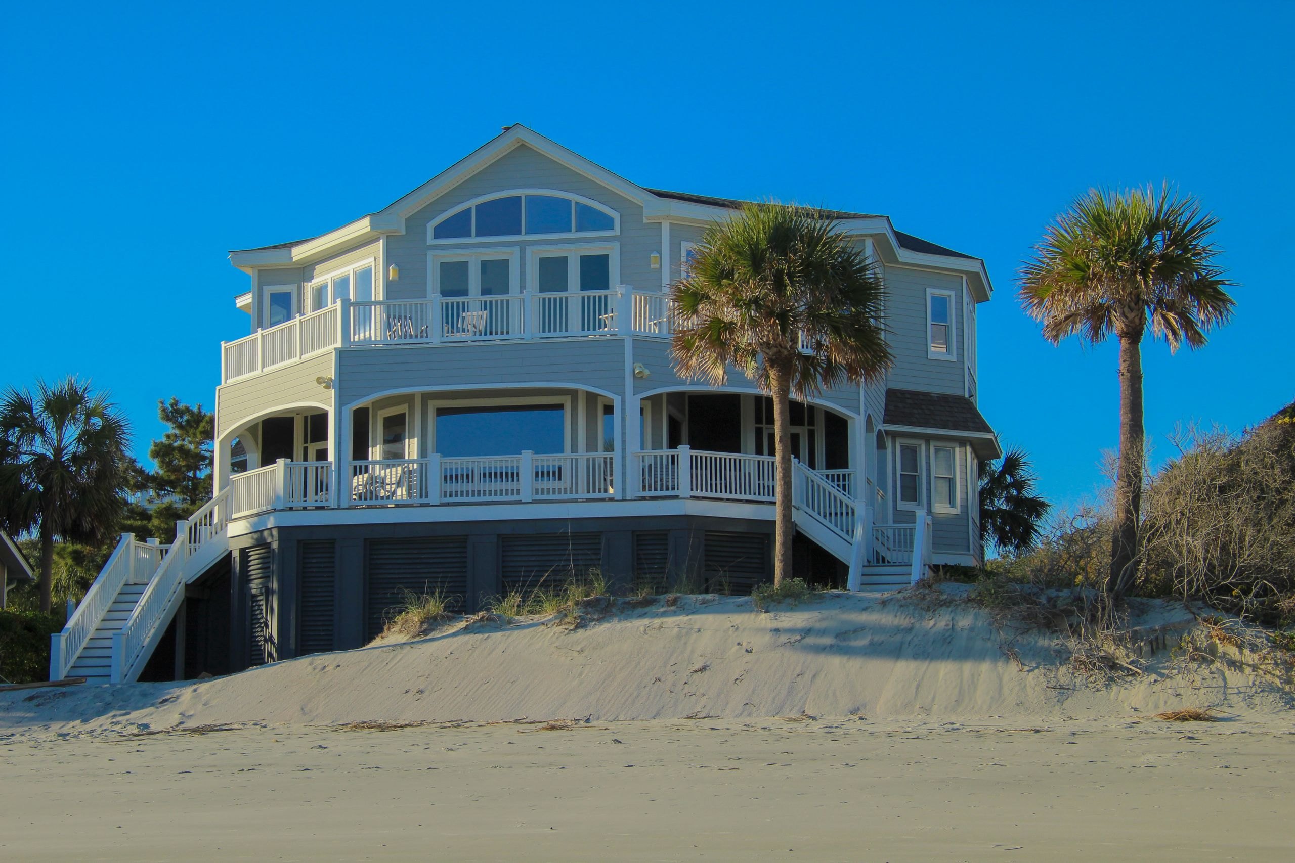 An elevated coastal home with wraparound porches above the dunes, palms and beach in front